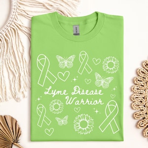 Puede incluir: Una camiseta verde lima con un diseño gráfico blanco. El diseño incluye lazos, mariposas, corazones y girasoles, junto con las palabras "Lyme Disease Warrior". La camiseta está hecha de un material suave.