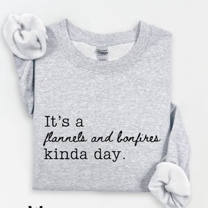 Puede incluir: Sudadera gris claro con el texto "It's a flannels and bonfires kinda day." La palabra "Ash" est&aacute; impresa debajo. La sudadera tiene cuello redondo y mangas largas.