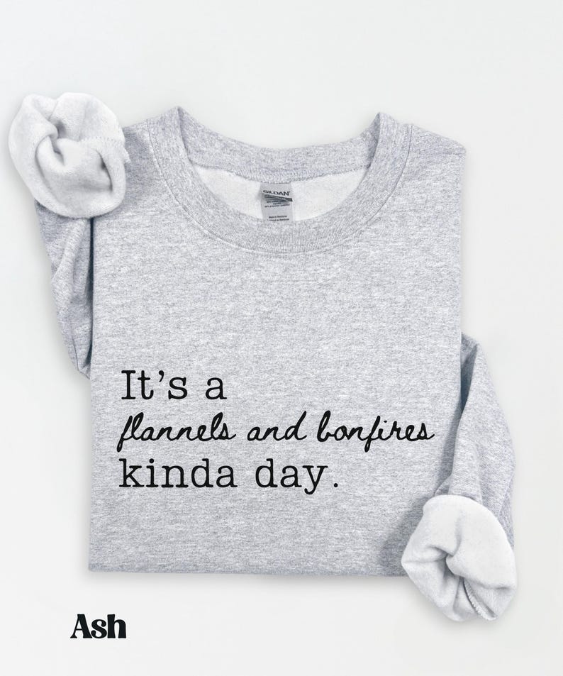 Puede incluir: Sudadera gris claro con el texto "It's a flannels and bonfires kinda day." La palabra "Ash" est&aacute; impresa debajo. La sudadera tiene cuello redondo y mangas largas.