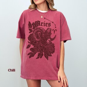 May include: A chili-coloured t-shirt with an Aries ram illustration and floral details. The design includes the word "Aries" and the Aries constellation. The shirt has a relaxed, oversized fit and is a deep red hue.