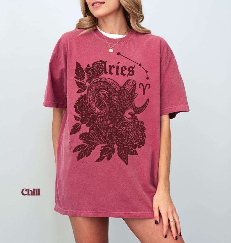 May include: A chili-coloured t-shirt with an Aries ram illustration and floral details. The design includes the word "Aries" and the Aries constellation. The shirt has a relaxed, oversized fit and is a deep red hue.