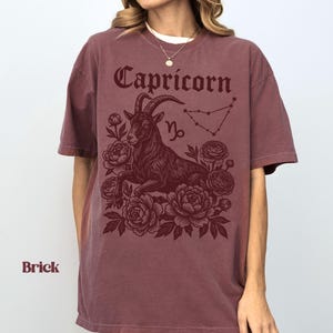 May include: Brick-colored t-shirt featuring a Capricorn design. The graphic includes the word "Capricorn" in a decorative font, a goat illustration, the Capricorn symbol, and a constellation outline. The design is surrounded by floral elements.