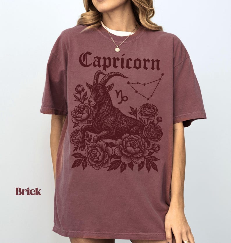 May include: Brick-colored t-shirt featuring a Capricorn design. The graphic includes the word "Capricorn" in a decorative font, a goat illustration, the Capricorn symbol, and a constellation outline. The design is surrounded by floral elements.
