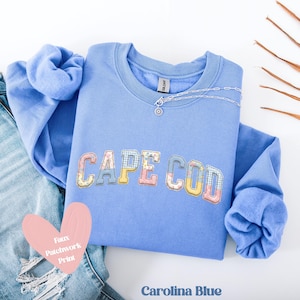Könnte beinhalten: Hellblaues Sweatshirt mit "CAPE COD" in einem Patchwork-Design. Eine silberne Halskette liegt auf dem Ausschnitt. Die Worte "Faux Patchwork Print" und "Carolina Blue" sind ebenfalls sichtbar. Das Sweatshirt ist gefaltet.