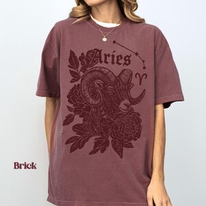 May include: Brick-coloured t-shirt with an Aries ram illustration, floral details, and the word "Aries". The Aries constellation is shown above the ram. The t-shirt is a relaxed fit.