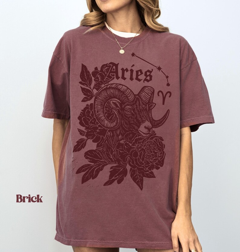 May include: Brick-coloured t-shirt with an Aries ram illustration, floral details, and the word "Aries". The Aries constellation is shown above the ram. The t-shirt is a relaxed fit.