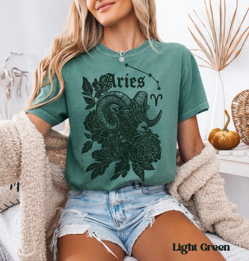 May include: Light green t-shirt with a black graphic of an Aries ram, flowers, and the word "Aries" in a decorative font. The Aries symbol is also included. The shirt is a short-sleeved, casual style.