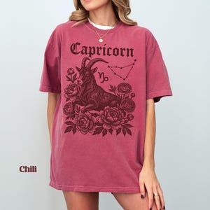 May include: A maroon t-shirt with the word "Capricorn" in a decorative font. The shirt features a detailed illustration of a goat surrounded by flowers and the Capricorn constellation. The word "Chili" is printed at the bottom.