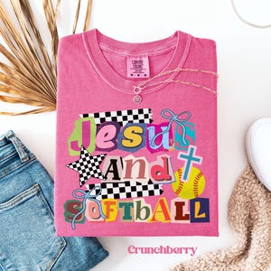 Può includere: Una maglietta rosa con le parole "Jesus and Softball" in un design colorato in stile collage. La maglietta presenta una stella, una croce e una grafica di softball. Una collana d'argento e jeans blu sono accanto alla maglietta.
