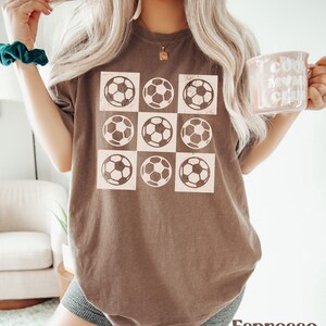 May include: A brown t-shirt with a graphic of nine white soccer balls arranged in a grid pattern. The shirt is a relaxed fit and has a distressed look.