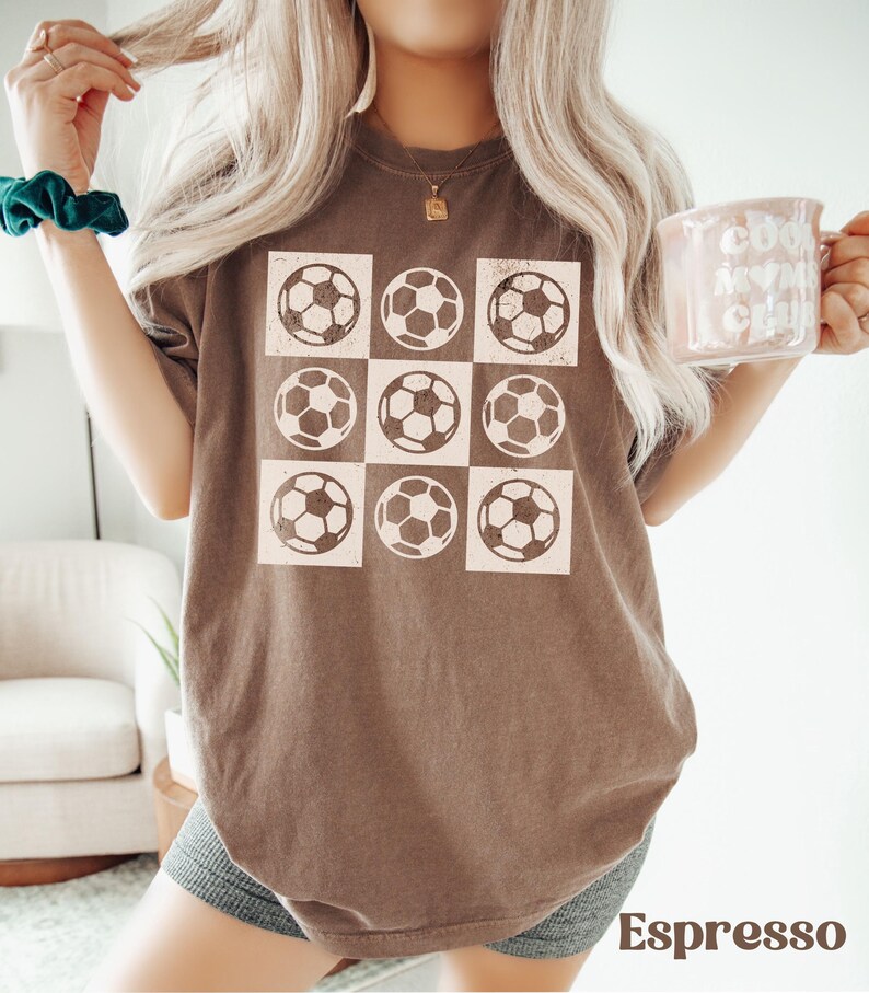 May include: A brown t-shirt with a graphic of nine white soccer balls arranged in a grid pattern. The shirt is a relaxed fit and has a distressed look.
