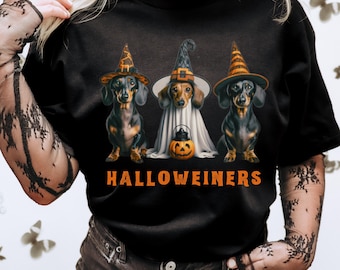 Op de afbeelding: Zwart T-shirt met drie teckels verkleed voor Halloween. De honden dragen heksenhoeden en een spookkostuum, met een pompoenlantaarn. De oranje tekst luidt "Halloweiners".