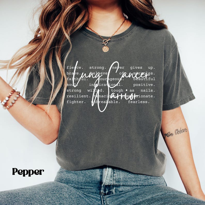 May include: A dark gray t-shirt with the text "Lung Cancer Warrior" printed in white. The text is defined as "fierce, strong, never gives up, brave, believe in miracles, faithful, courageous, beautiful soul, inspirational, positive, strong willed, tough as nails, resilient, tenacious, passionate, fighter, unbreakable, fearless".
