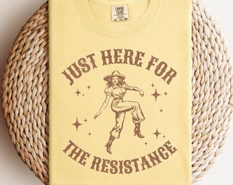 Op de afbeelding: Geel T-shirt met bruine tekst die "Just Here For The Resistance" luidt. Een cartoonvrouw in een cowboyhoed en laarzen danst in het midden van de tekst.