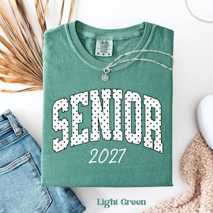 Peut inclure: Un t-shirt vert clair Comfort Colors avec le mot "SENIOR" en lettres &agrave; pois blancs et l'ann&eacute;e "2027" en blanc. Un collier argent&eacute; avec un pendentif est sur le t-shirt. Les mots "Light Green" sont en bas.