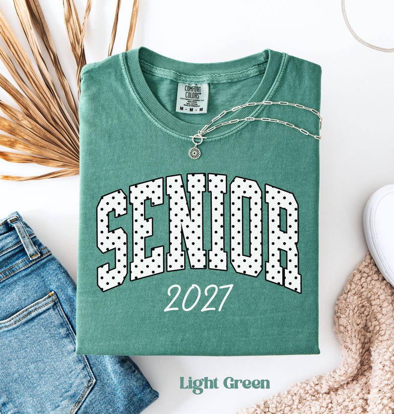 Peut inclure: Un t-shirt vert clair Comfort Colors avec le mot "SENIOR" en lettres &agrave; pois blancs et l'ann&eacute;e "2027" en blanc. Un collier argent&eacute; avec un pendentif est sur le t-shirt. Les mots "Light Green" sont en bas.
