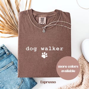 May include: Espresso-colored t-shirt with the words "dog walker" and a paw print graphic. A silver necklace with a sun pendant rests on the shirt. The shirt is folded, with blue jeans and a white shoe visible.