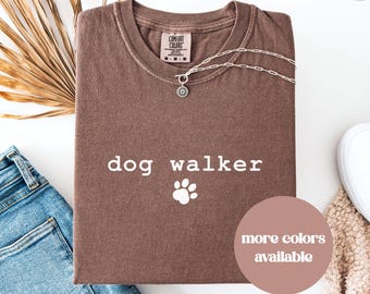 May include: Espresso-colored t-shirt with the words "dog walker" and a paw print graphic. A silver necklace with a sun pendant rests on the shirt. The shirt is folded, with blue jeans and a white shoe visible.