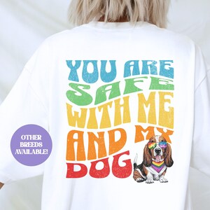 May include: White t-shirt with a rainbow-colored graphic that says "You are safe with me and my dog". A cartoon basset hound wearing rainbow sunglasses is featured on the shirt.