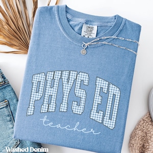 May include: A washed denim blue crewneck t-shirt featuring the text PHYS ED in a light blue and white gingham pattern, with the word teacher written below in white script. A silver chain necklace with a circular pendant rests on the neckline.