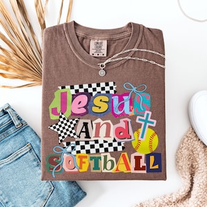Può includere: Una maglietta marrone Comfort Colors con una grafica colorata che dice "Jesus And Softball". Il design include una croce, una palla da softball e una stella con un motivo a scacchi. Una collana d'argento è drappeggiata sulla maglietta.