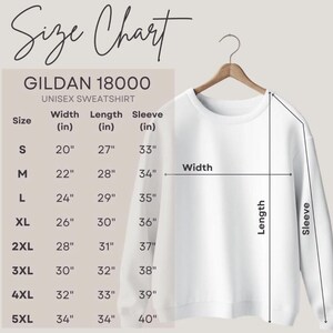 Puede incluir: Una sudadera unisex blanca Gildan 18000 cuelga de una percha de madera. La imagen incluye una tabla de tallas con medidas en cm para el ancho, largo y largo de manga, que van desde la talla S hasta la 5XL.