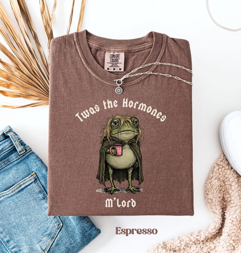 May include: A brown t-shirt featuring a graphic of a green frog with long hair wearing a dark cloak and holding a pink mug. The text above the frog reads "Twas the Hormones" and the text below reads "M'lord". The shirt has a Comfort Colors label.