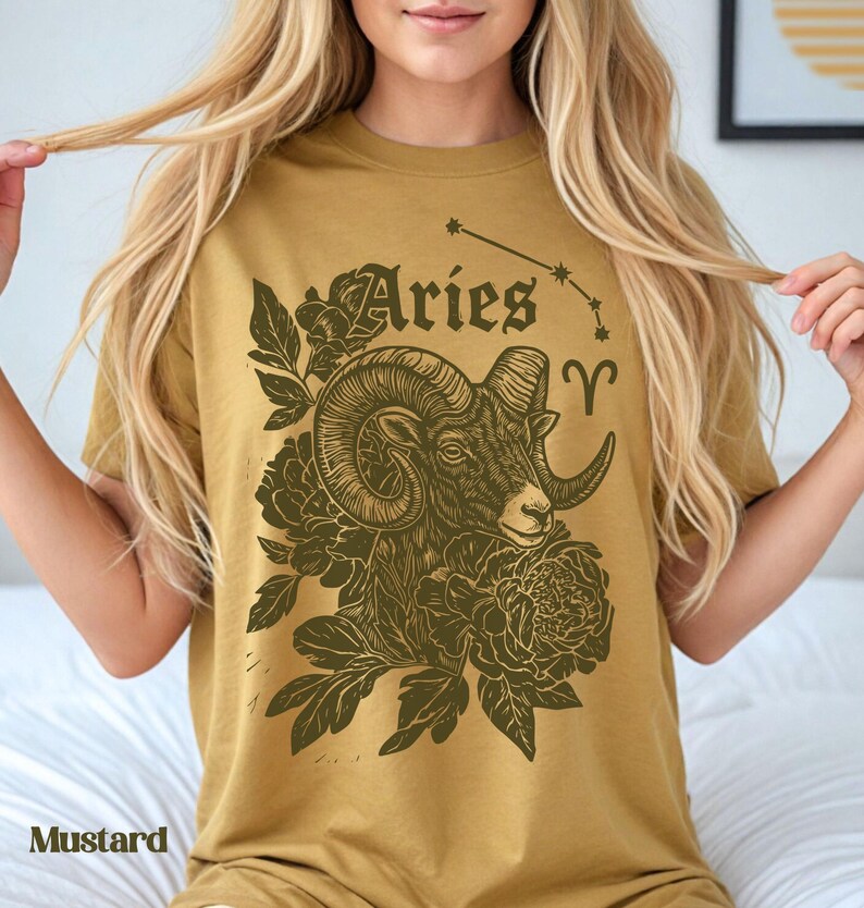 May include: Mustard-coloured t-shirt with a detailed illustration of an Aries ram's head, floral elements, and the word "Aries". The design includes the Aries constellation symbol. The t-shirt is a mustard colour.