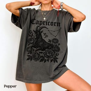 May include: Dark gray t-shirt featuring a black Capricorn graphic. The design includes a goat, floral elements, the word "Capricorn", and the Capricorn constellation. The shirt has a relaxed fit.