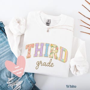 Op de afbeelding: Wit sweatshirt met de tekst "THIRD grade" in een kleurrijk patchwork-ontwerp. Het woord "THIRD" heeft bloemen-, ruit- en streepmotieven. Een delicate ketting en een roze hartvormig label zijn ook zichtbaar.