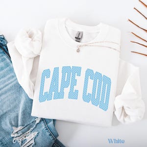Könnte beinhalten: Ein weißes Sweatshirt mit dem Text "CAPE COD" in hellblauen, gepunkteten Buchstaben. Eine silberne Halskette mit einem kleinen Anhänger liegt auf dem Sweatshirt. Das Wort "White" befindet sich am unteren Bildrand.