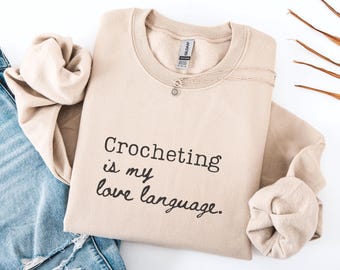 May include: Sand-colored sweatshirt with the text "Crocheting is my love language" in black script. A silver necklace with a small pendant is on the front. The sweatshirt is folded with a pair of blue jeans.