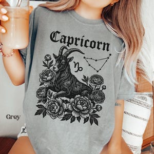 May include: Grey t-shirt featuring a black Capricorn zodiac design. The design includes a goat, flowers, the Capricorn symbol, and the word "Capricorn" in a decorative font. The shirt is a soft, comfortable material.