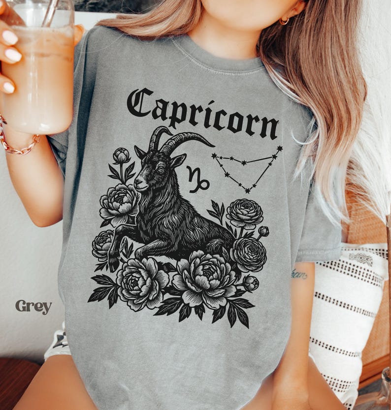 May include: Grey t-shirt featuring a black Capricorn zodiac design. The design includes a goat, flowers, the Capricorn symbol, and the word "Capricorn" in a decorative font. The shirt is a soft, comfortable material.