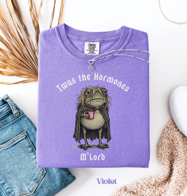 May include: A violet t-shirt featuring a graphic of a frog with long hair wearing a dark cape and holding a pink mug. The text above the frog reads 'Twas the Hormones' and the text below reads 'M'lord'. The shirt has a crew neck and short sleeves.