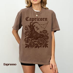 May include: Espresso-colored t-shirt featuring a Capricorn zodiac design. The graphic includes the word "Capricorn" in a decorative font, a goat illustration, constellation, and floral elements. The shirt is a relaxed fit.
