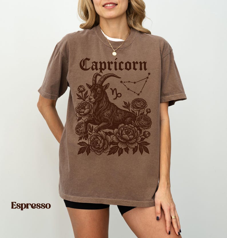 May include: Espresso-colored t-shirt featuring a Capricorn zodiac design. The graphic includes the word "Capricorn" in a decorative font, a goat illustration, constellation, and floral elements. The shirt is a relaxed fit.