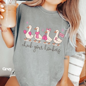 Pu&ograve; includere: T-shirt grigia con un'illustrazione di quattro anatre che camminano in fila. Ogni anatra indossa accessori rosa, come guantoni da boxe e una sciarpa. Sotto le anatre c'&egrave; la scritta "check your honkers".
