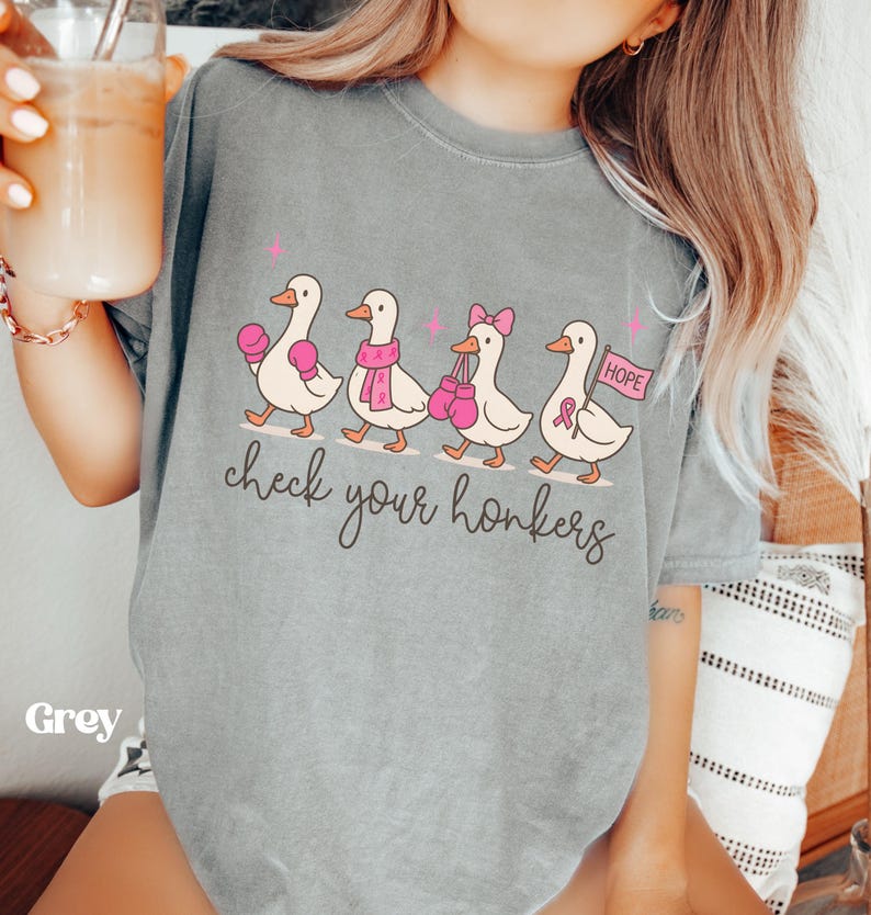 Pu&ograve; includere: T-shirt grigia con un'illustrazione di quattro anatre che camminano in fila. Ogni anatra indossa accessori rosa, come guantoni da boxe e una sciarpa. Sotto le anatre c'&egrave; la scritta "check your honkers".