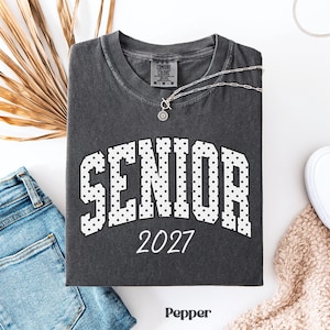 Peut inclure: Un t-shirt gris fonc&eacute; avec le mot "SENIOR" en blanc, lettres pointill&eacute;es et l'ann&eacute;e "2027" en blanc. Un collier argent&eacute; avec un pendentif est sur le t-shirt. Le t-shirt est pli&eacute; sur une surface blanche &agrave; c&ocirc;t&eacute; d'un jean bleu et d'une chaussure blanche.