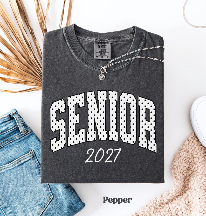 Peut inclure: Un t-shirt gris fonc&eacute; avec le mot "SENIOR" en blanc, lettres pointill&eacute;es et l'ann&eacute;e "2027" en blanc. Un collier argent&eacute; avec un pendentif est sur le t-shirt. Le t-shirt est pli&eacute; sur une surface blanche &agrave; c&ocirc;t&eacute; d'un jean bleu et d'une chaussure blanche.