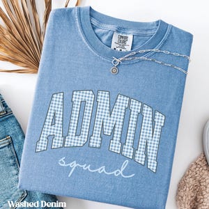 May include: A washed denim blue short-sleeve t-shirt featuring the word ADMIN in a light blue and white gingham block font, with the word squad written below in a white cursive script. A silver chain necklace rests on the collar area.