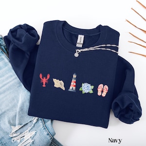 May include: Navy blue sweatshirt with coastal-themed illustrations. Features a red lobster, seashell, lighthouse, blue hydrangea, and flip-flops. A silver necklace with a pendant is draped across the sweatshirt. The word "Navy" is printed at the bottom.