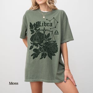 Op de afbeelding: Een mosgroen t-shirt met een Weegschaal sterrenbeeld ontwerp. De afbeelding bevat het woord "Libra", een weegschaal, bloemmotieven en het Weegschaal sterrenbeeld. Het t-shirt heeft een relaxte pasvorm.