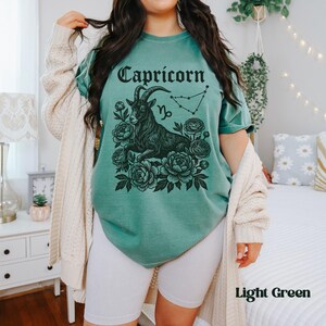 May include: Light green t-shirt featuring a black Capricorn graphic with a goat, flowers, and constellation. The word "Capricorn" is arched across the top. The shirt is a light green color.