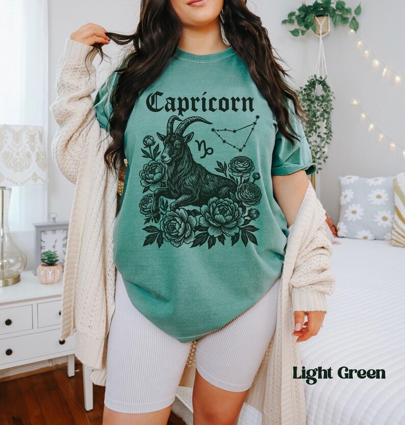 May include: Light green t-shirt featuring a black Capricorn graphic with a goat, flowers, and constellation. The word "Capricorn" is arched across the top. The shirt is a light green color.