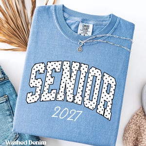 Peut inclure: Un t-shirt bleu clair "Washed Denim" avec le mot "SENIOR" en grandes lettres bord&eacute;es de noir et remplies de pois blancs. L'ann&eacute;e "2027" est imprim&eacute;e en dessous en blanc. Un collier argent&eacute; avec un pendentif est visible.
