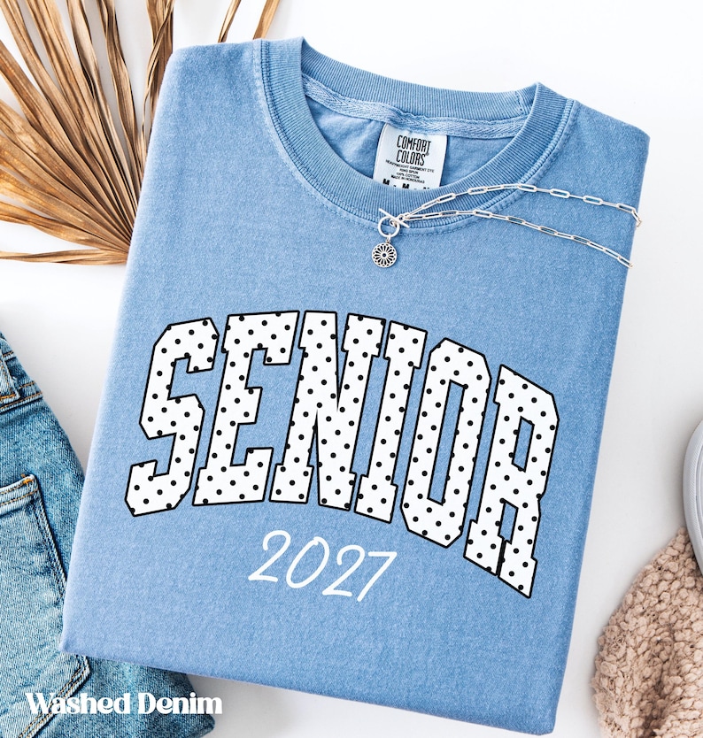 Peut inclure: Un t-shirt bleu clair "Washed Denim" avec le mot "SENIOR" en grandes lettres bord&eacute;es de noir et remplies de pois blancs. L'ann&eacute;e "2027" est imprim&eacute;e en dessous en blanc. Un collier argent&eacute; avec un pendentif est visible.