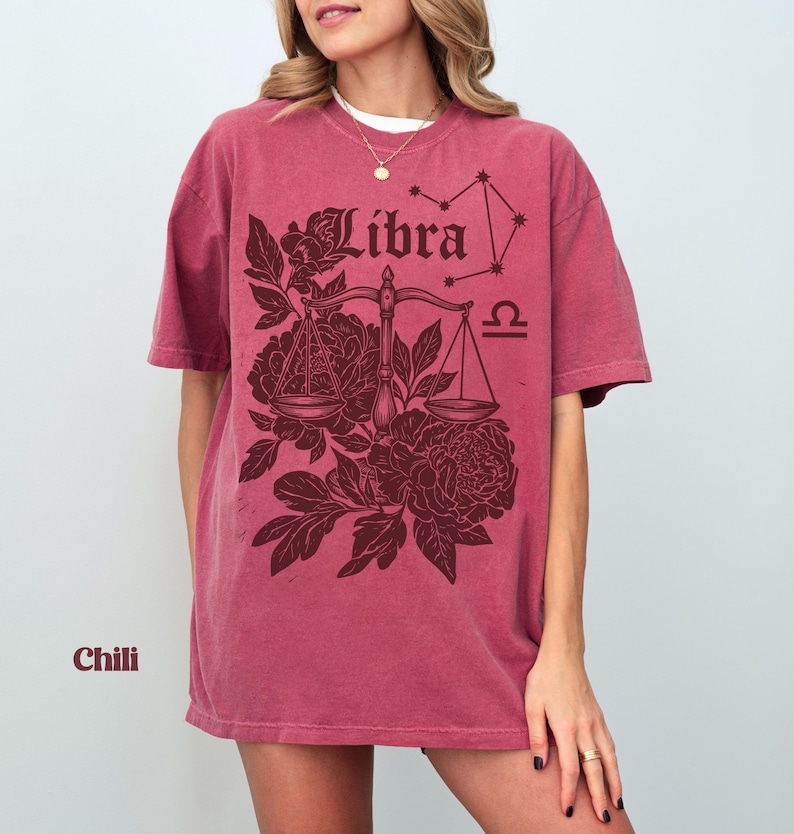 Op de afbeelding: Een chili-kleurig T-shirt met korte mouwen met een Weegschaal-sterrenbeeldontwerp. Het ontwerp bevat het woord "Libra" in een decoratief lettertype, een weegschaal, bloemelementen en het Weegschaal-sterrenbeeld.