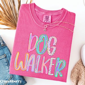 May include: A pink short-sleeved t-shirt with a crew neckline featuring the text DOG WALKER in a multi-coloured, marbled, and speckled graphic print. The shirt has a Comfort Colors brand label at the inner neck and a gold-toned chain necklace.